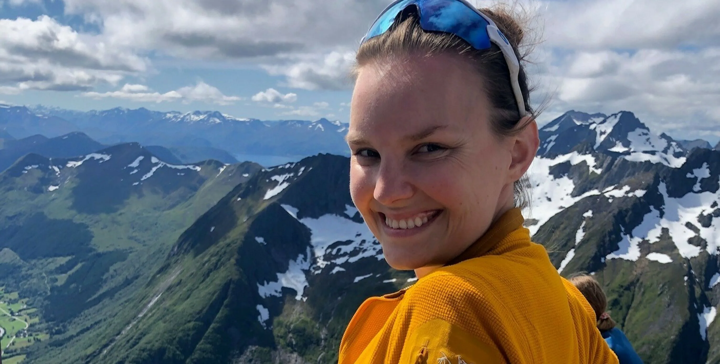 Smiling hiker in a yellow jacket on a mountaintop, overlooking dramatic Sunnmøre mountains with patches of snow.