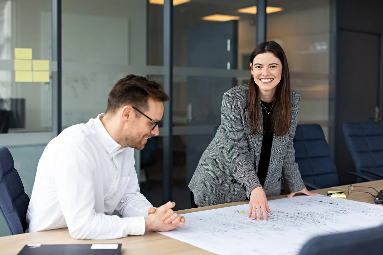 Two VARD colleagues smiling while reviewing ship design drawings spread across a meeting table in a modern office.