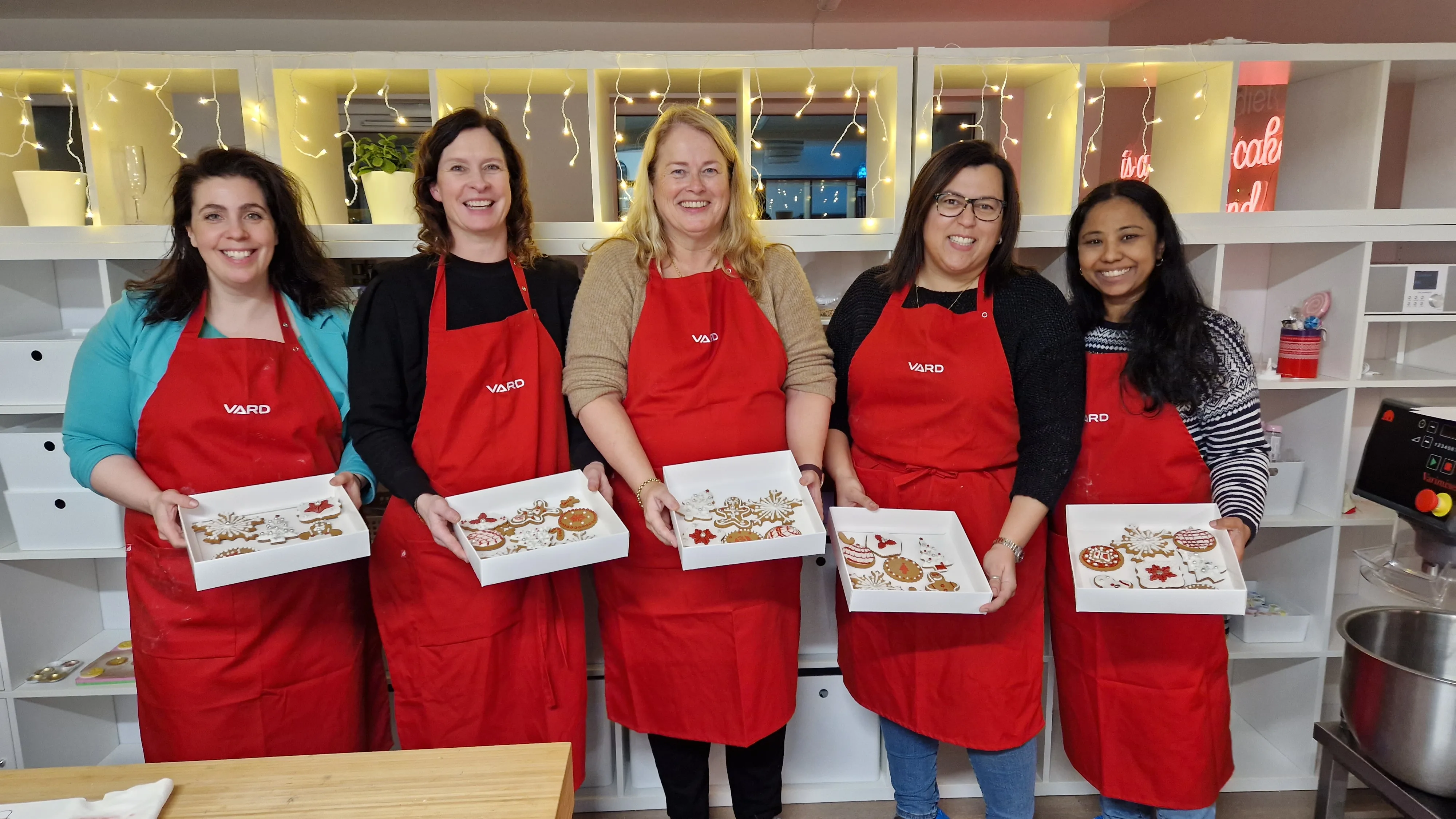 Five VARD employees wearing red VARD aprons, smiling and holding trays of freshly decorated cookies in a kitchen.