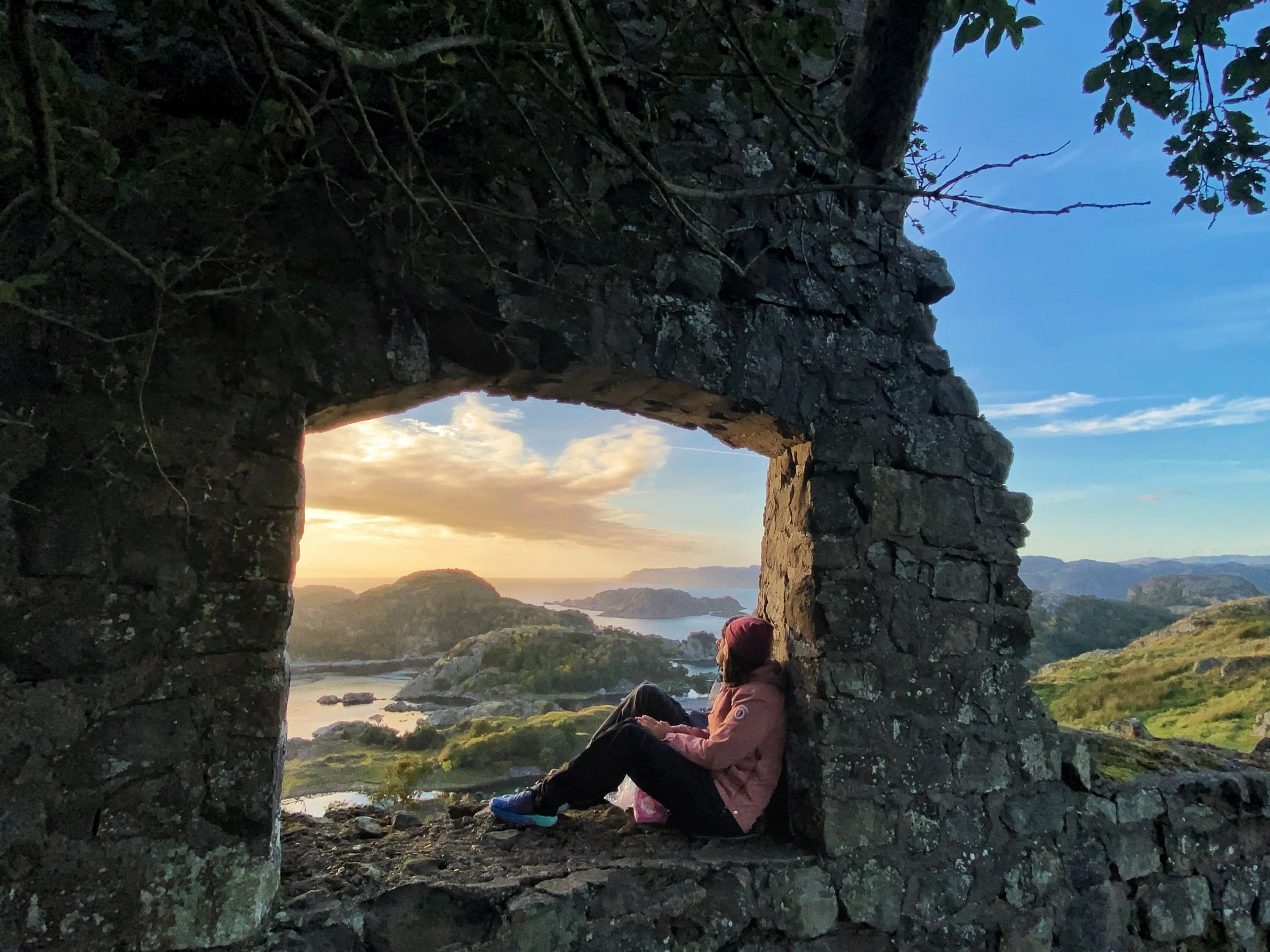 A lady sitting in an opening in an old stone house with a beautiful view