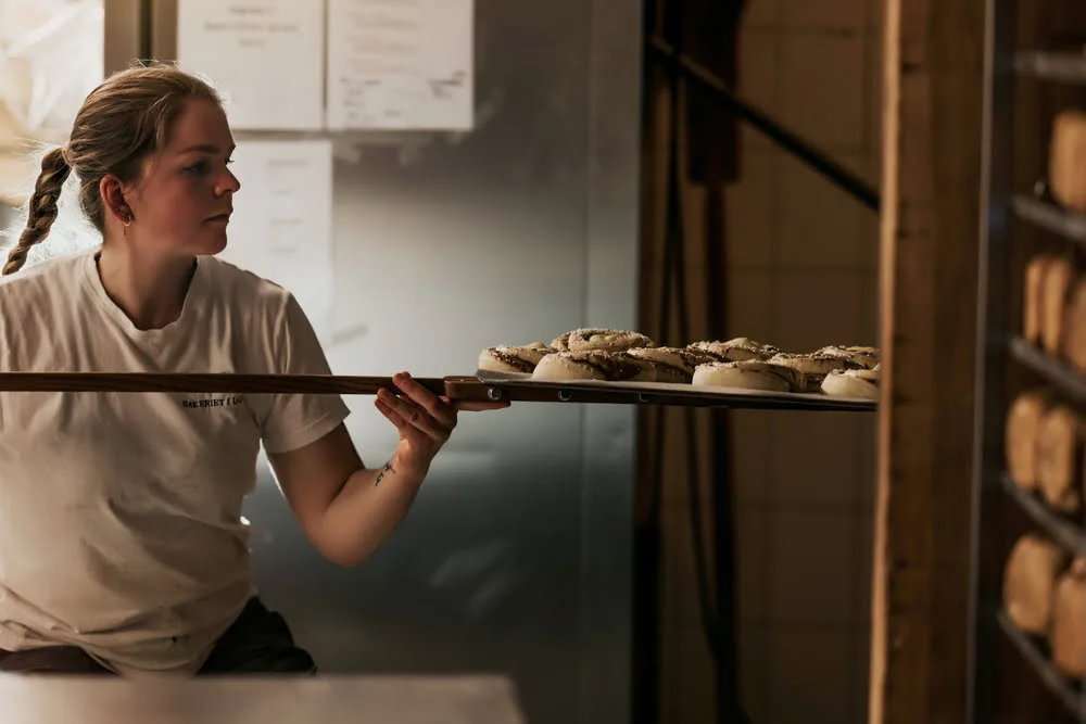 A baker at Bakeriet i Lom holding a wooden peel with freshly baked cinnamon buns, ready for the oven