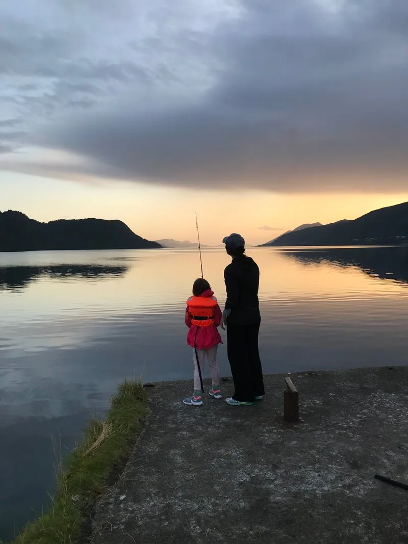 An adult and a child stand with a fishing rod on a small pier, looking out over a calm fjord at sunset
