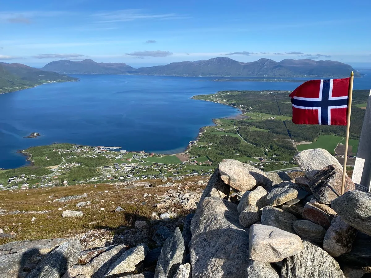 Norwegian flag on a rocky summit overlooking Tomrefjord, the village and surrounding mountains in bright sunshine