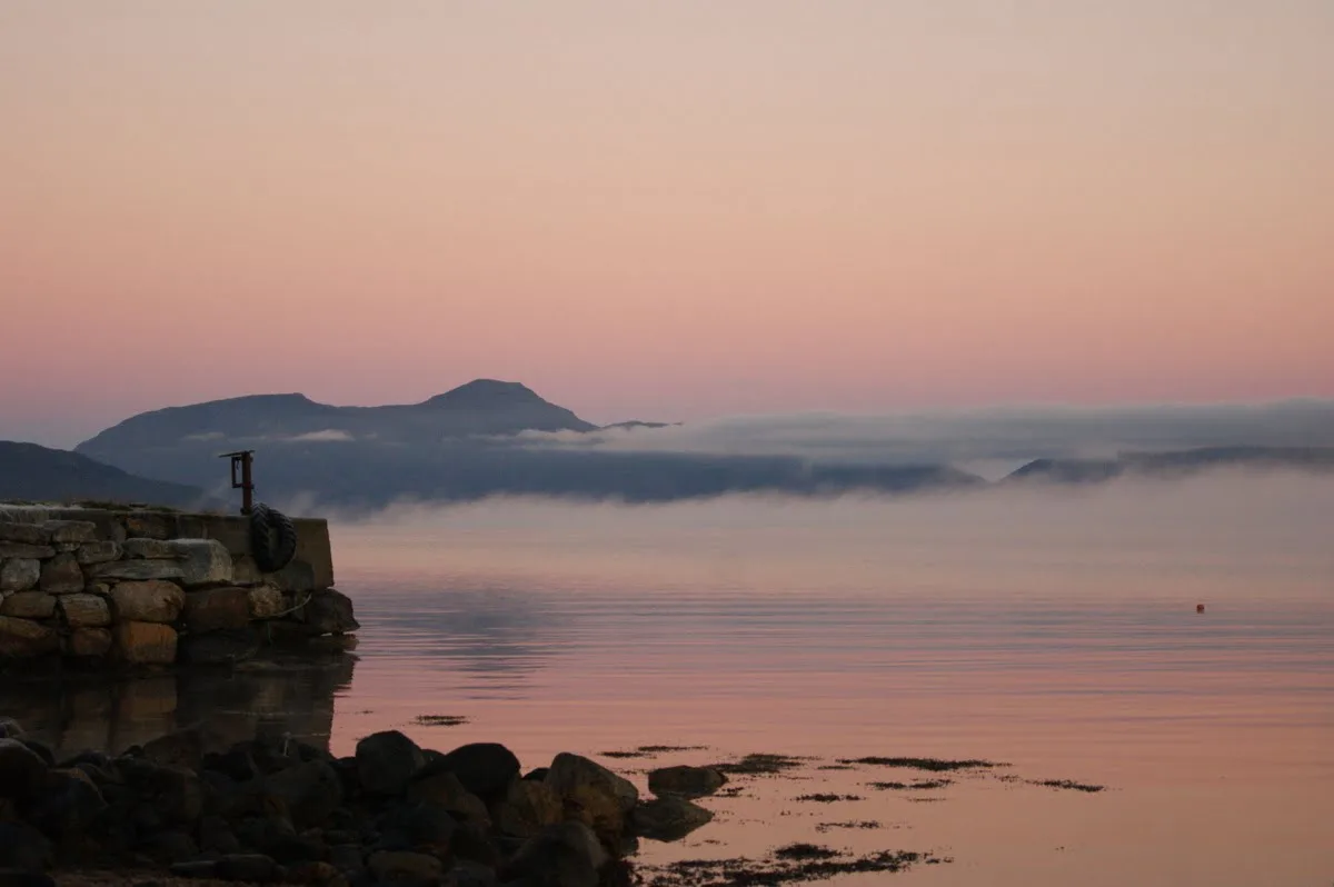 Calm evening at Tomrefjord with a stone pier in the foreground, misty mountains and a pink sky reflected in the water