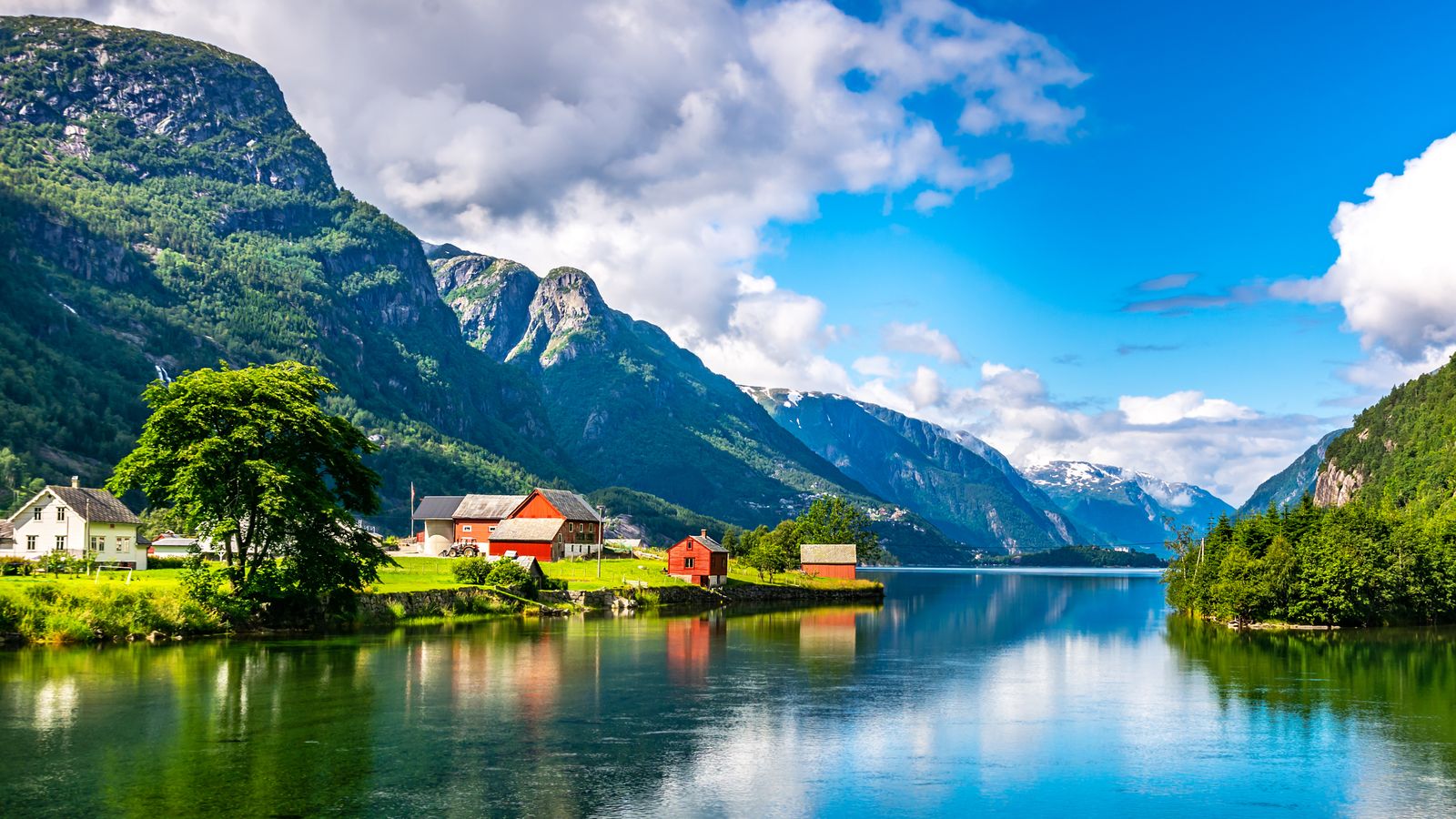 Red wooden houses by a calm fjord in western Norway, with steep green mountains and snow-capped peaks in the background